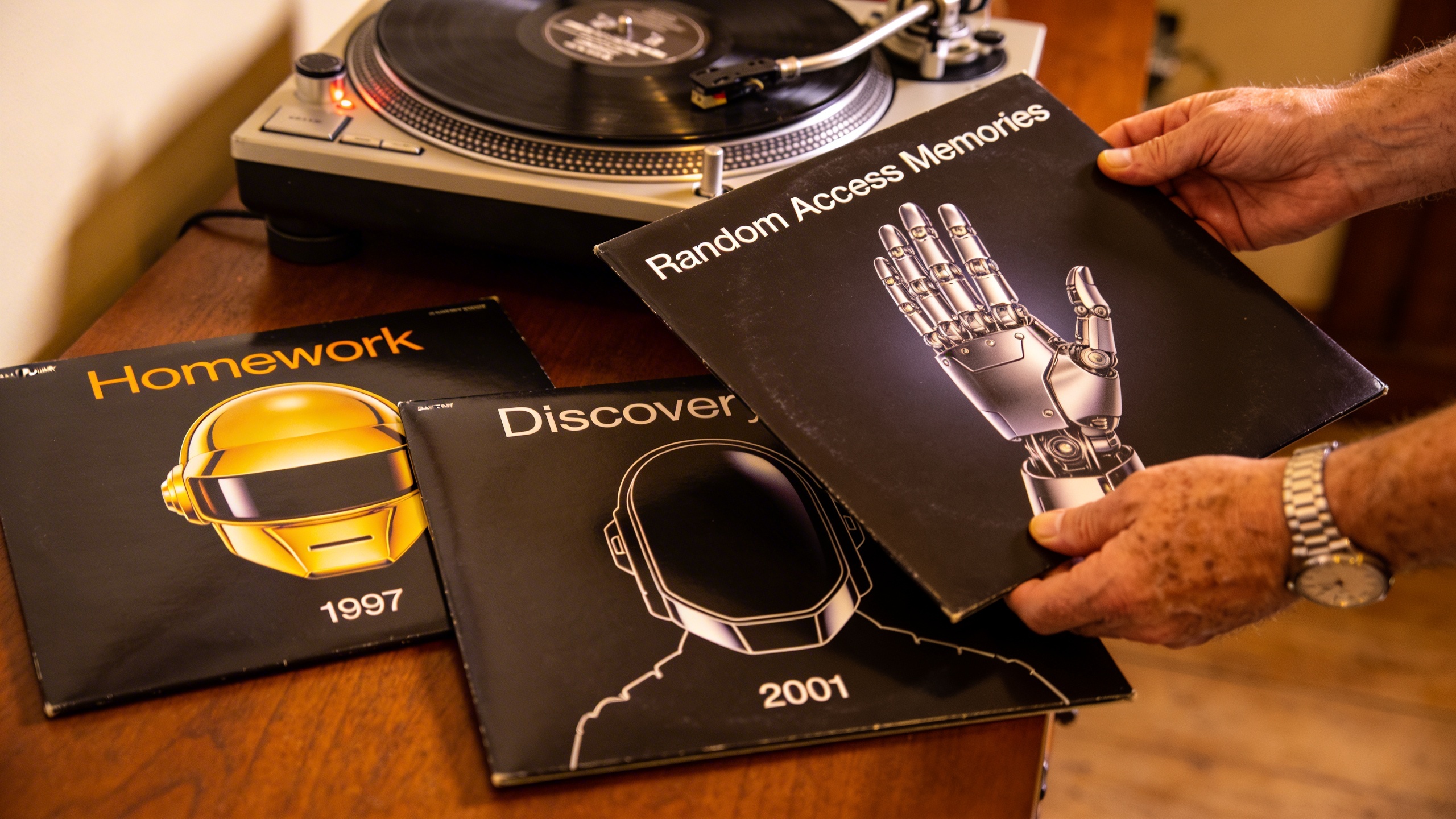 Photorealistic: Collection of essential Daft Punk albums arranged on rich mahogany surface - Homework, Discovery, and Random Access Memories vinyl covers displayed with warm tungsten lighting, vintage